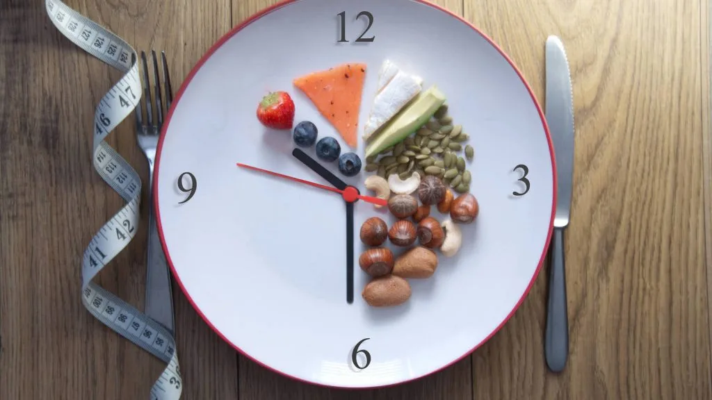 Reloj de plato con alimentos saludables representando el ayuno intermitente, acompañado de cubiertos y cinta métrica sobre una mesa de madera